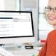 Woman in red blouse smiling in front of a desk with computer showing a screenshot of GoldMine CRM