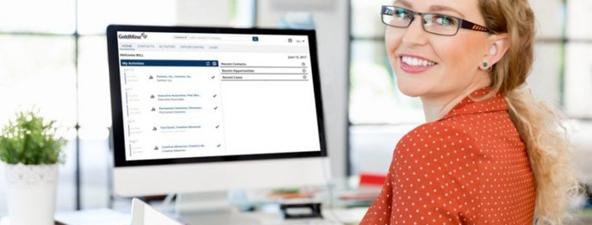 Woman in red blouse smiling in front of a desk with computer showing a screenshot of GoldMine CRM