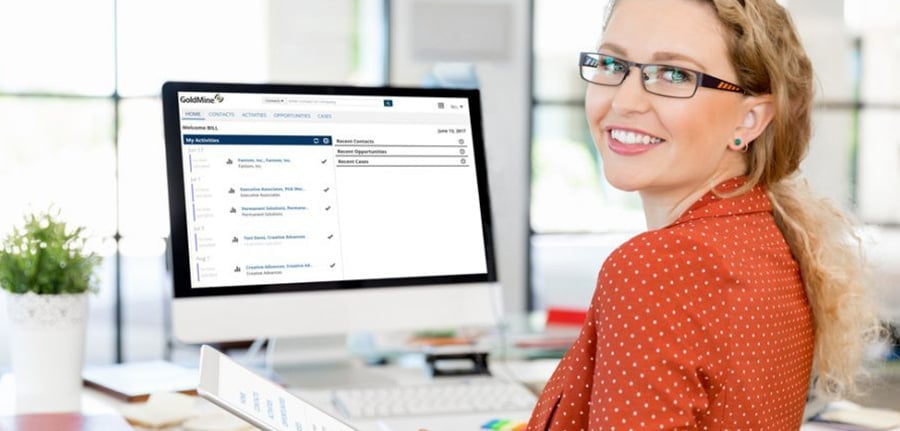 Woman in red blouse smiling in front of a desk with computer showing a screenshot of GoldMine CRM