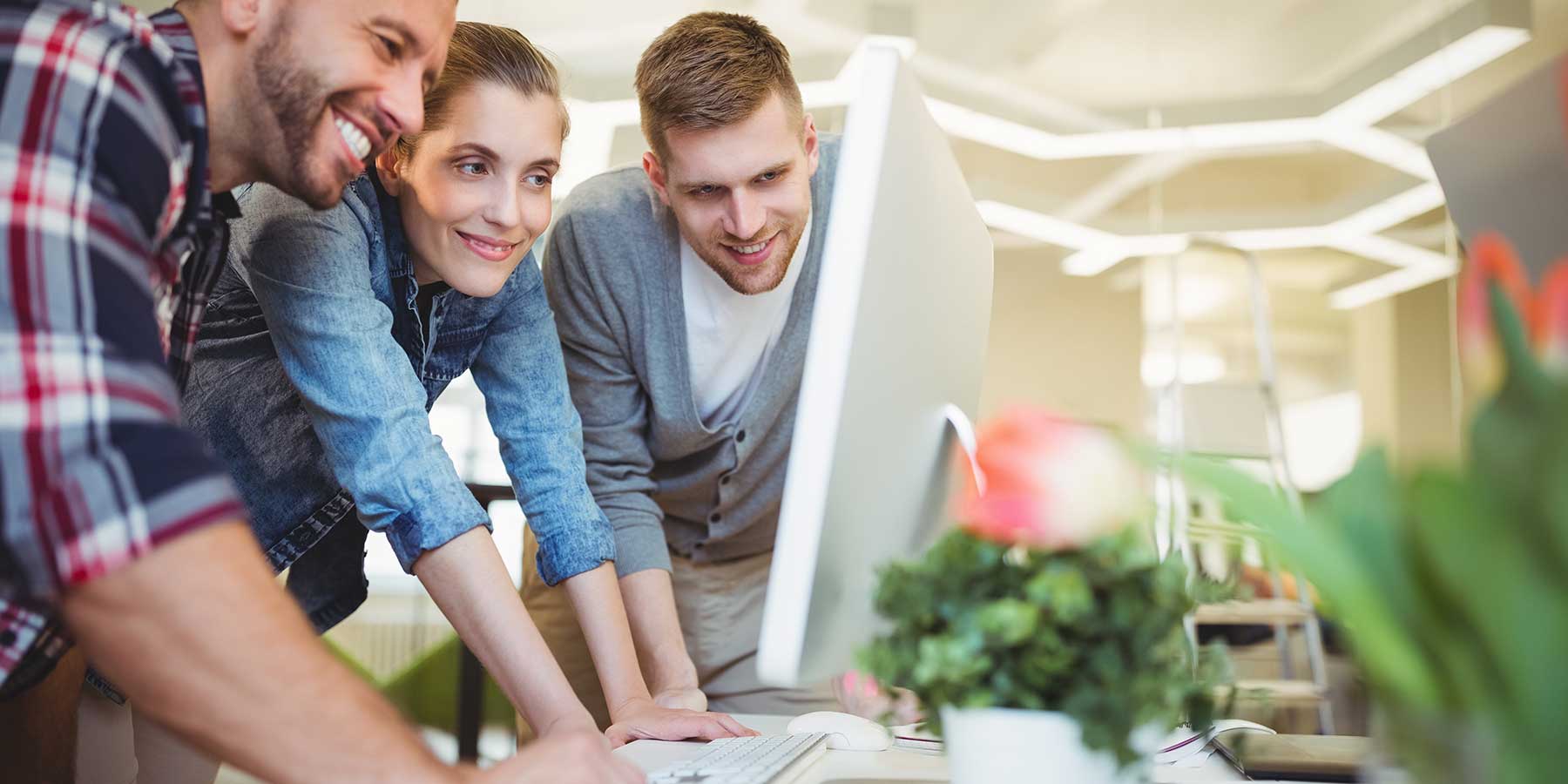 a woman and two men all in their twenties crowded around a desktop looking at the screen and smiling